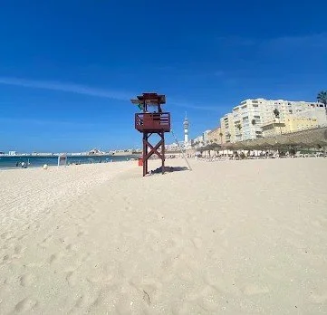 Empty sandy beach with a lifeguard tower, ocean, and buildings in the background under a clear blue sky.