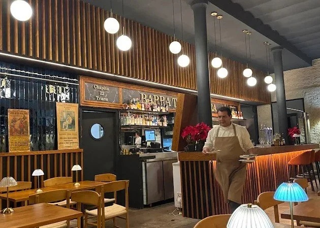Inside a restaurant with wooden decor, hanging globe lights, a bar area, and a server preparing a table.
