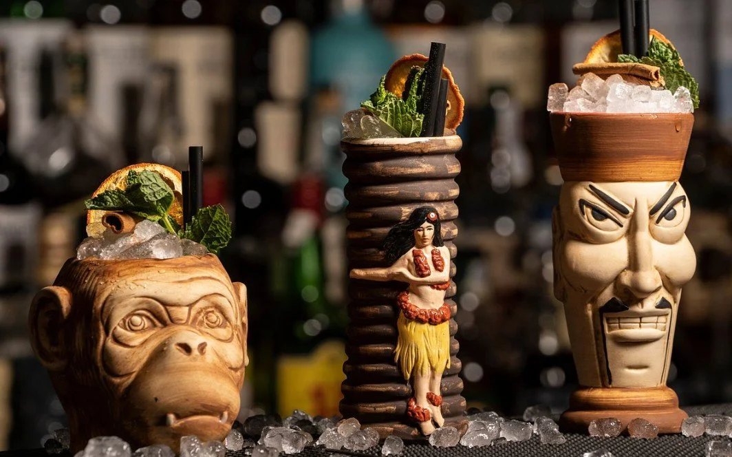 Three tiki-style mugs with drinks, garnished with mint, lemon, and other citrus slices, placed on ice. The mugs have unique carved faces and are set against a blurred bar background.