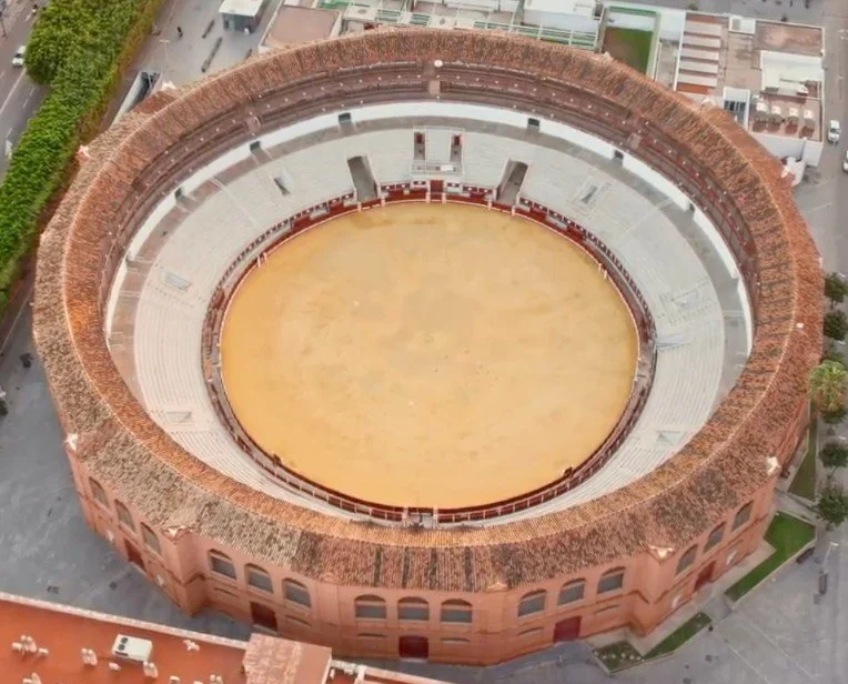 Aerial view of a large, round bullring with red brick exterior and a sandy arena in the center.