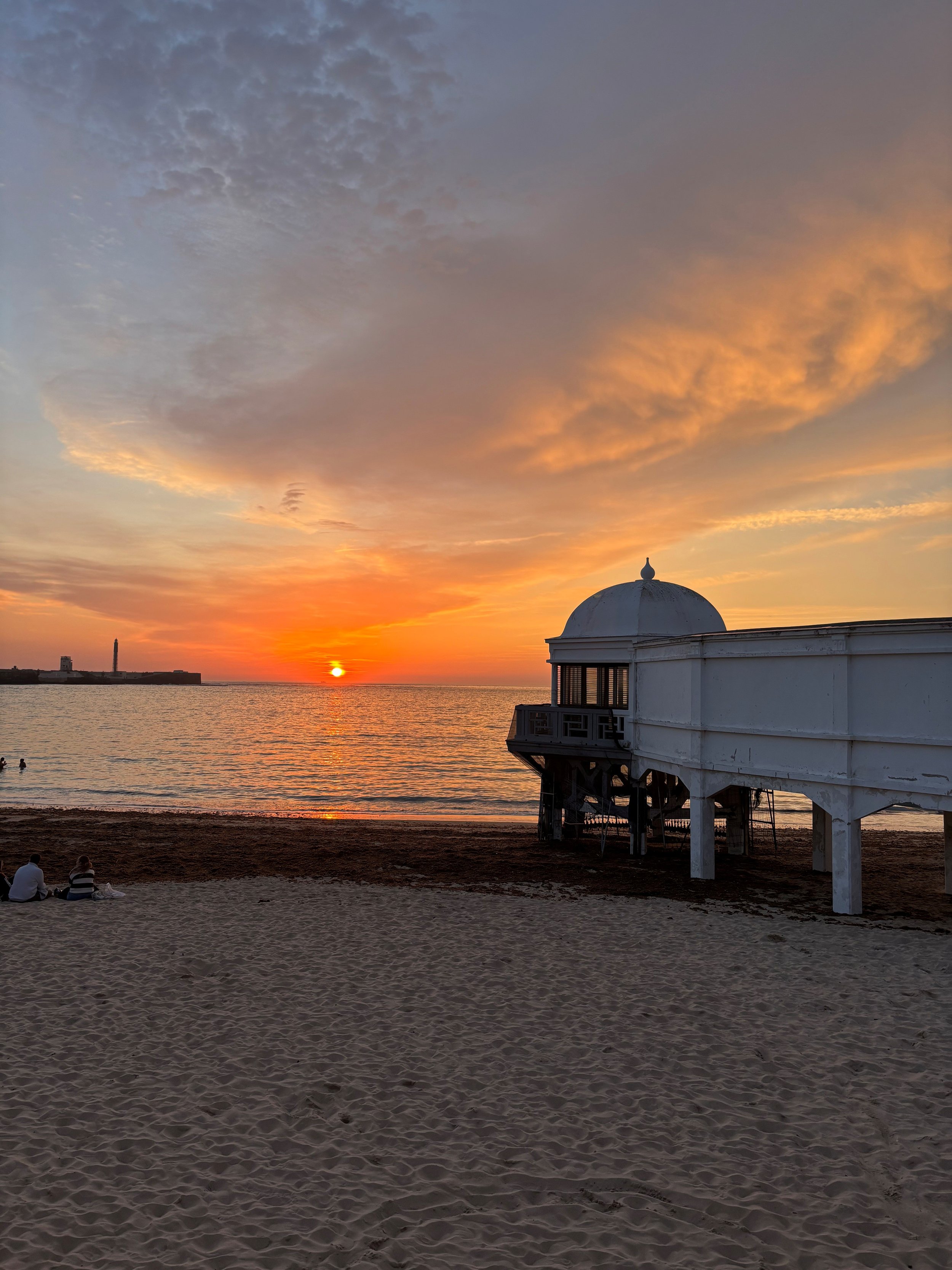 A sunset over the ocean with a large cloud in the sky, a white pier extending into the water, and a sandy beach with a few people sitting and walking.