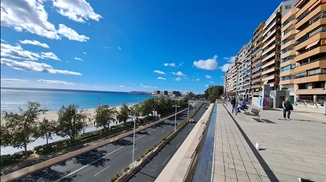 A seaside promenade with a paved walkway, benches, and people walking, alongside multi-story apartment buildings, with a view of a beach, the ocean, and a partly cloudy blue sky.