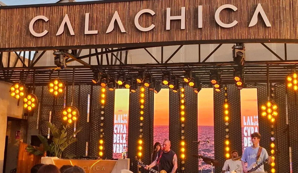 A stage with a large sign reading 'CALACHA' at the top, decorated with orange yellow lights and screens showing a sunset over the ocean. Live musicians are performing.