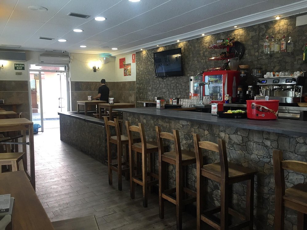 Interior of a small cafe with a stone countertop bar, wooden chairs, a wall-mounted TV, and a drink station with a red coffee machine and shelves with bottles.