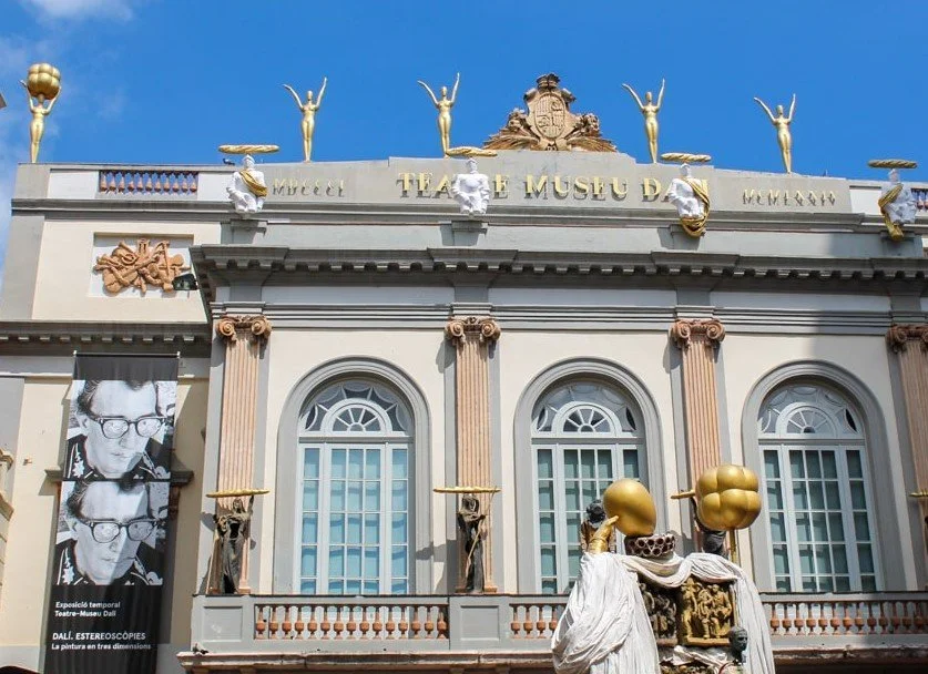 Facade of the Teatro Museo Dalí with classical architectural features, statues on the roof, and promotional banner with a black-and-white photo of Dalí wearing glasses.