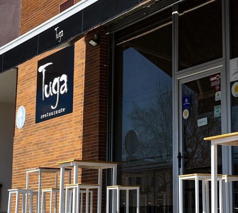 Exterior view of Tuga restaurant with outdoor seating and a large window reflecting trees.