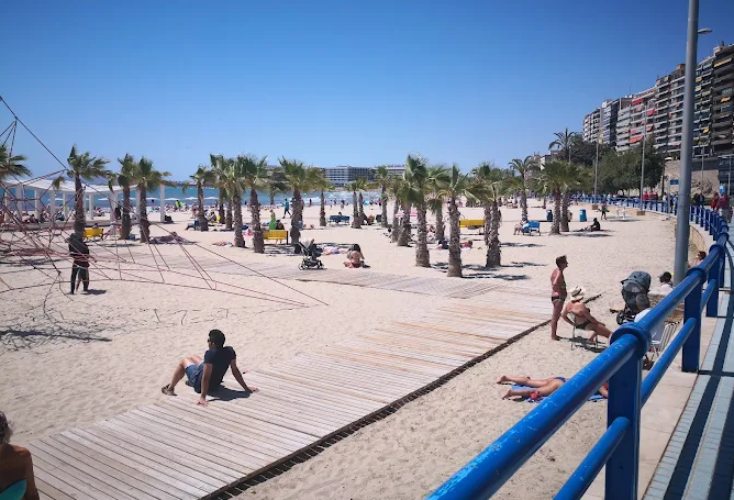 People relaxing on the sandy beach with palm trees, wooden walkway, and high-rise buildings in the background under a clear blue sky.