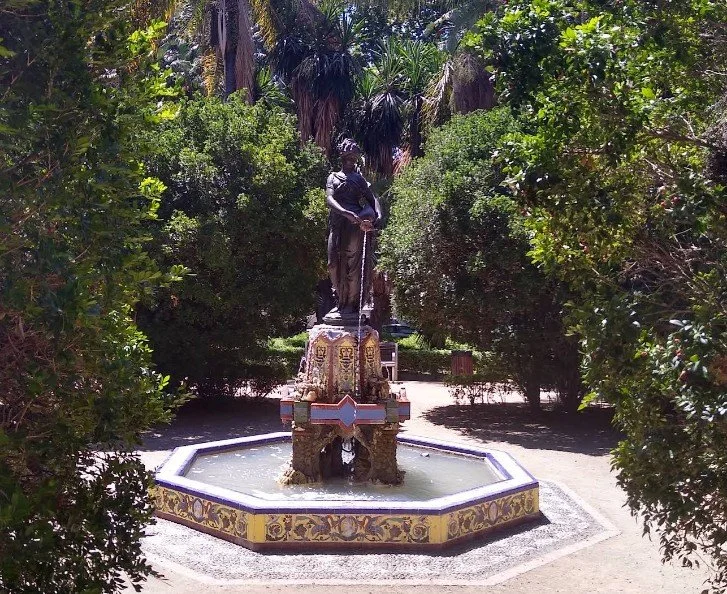A bronze statue of a man holding a sword stands in the center of a decorative fountain surrounded by lush green trees in a park.