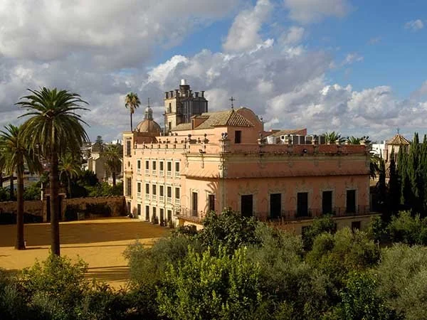 Historic pink building with Mediterranean architecture surrounded by trees and palm trees, under a partly cloudy sky.