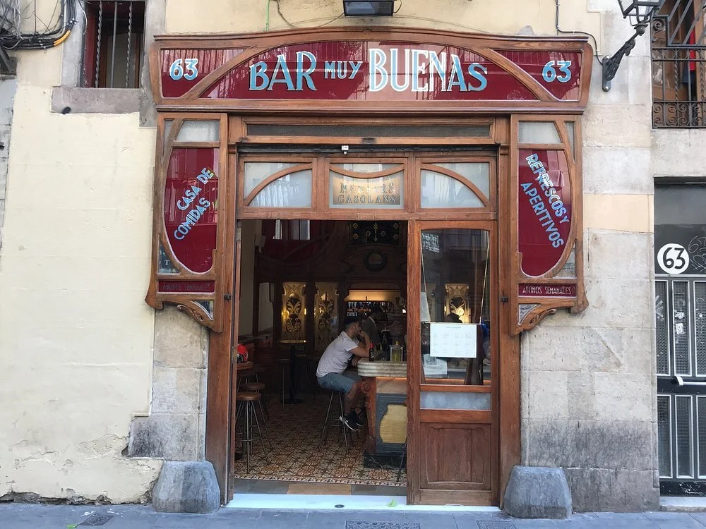 Exterior view of a bar called 'Bar Muy Buenas' with a wooden entrance and red signage, located at 63.