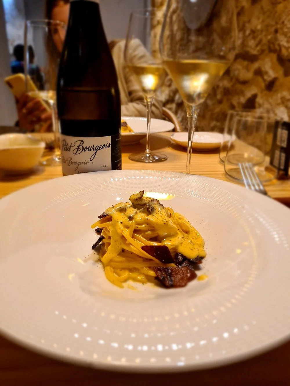 Plate of pasta with creamy sauce and bacon, set on a dining table with glasses of white wine and a bottle of wine in the background.