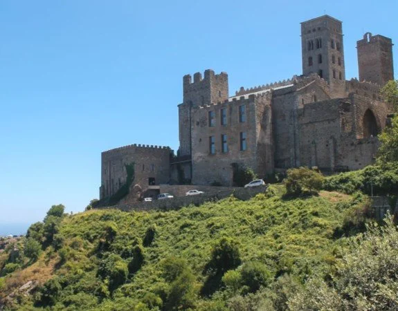 A large stone castle with multiple towers on a hill, with greenery and a few parked cars in front.