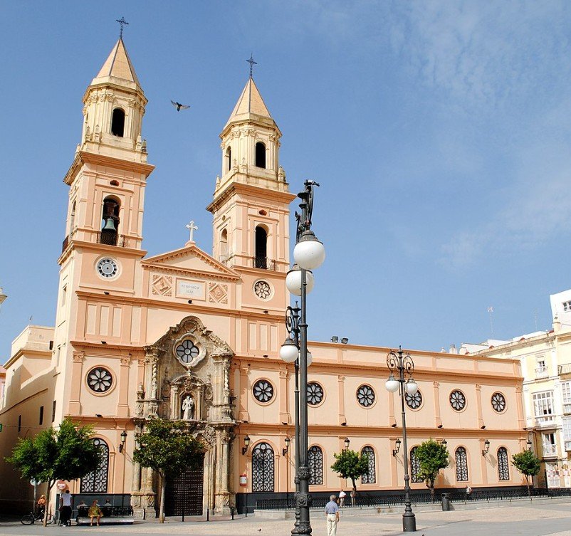Pink church with twin towers, decorative facade, arched windows, and several street lamps in front, under a blue sky.