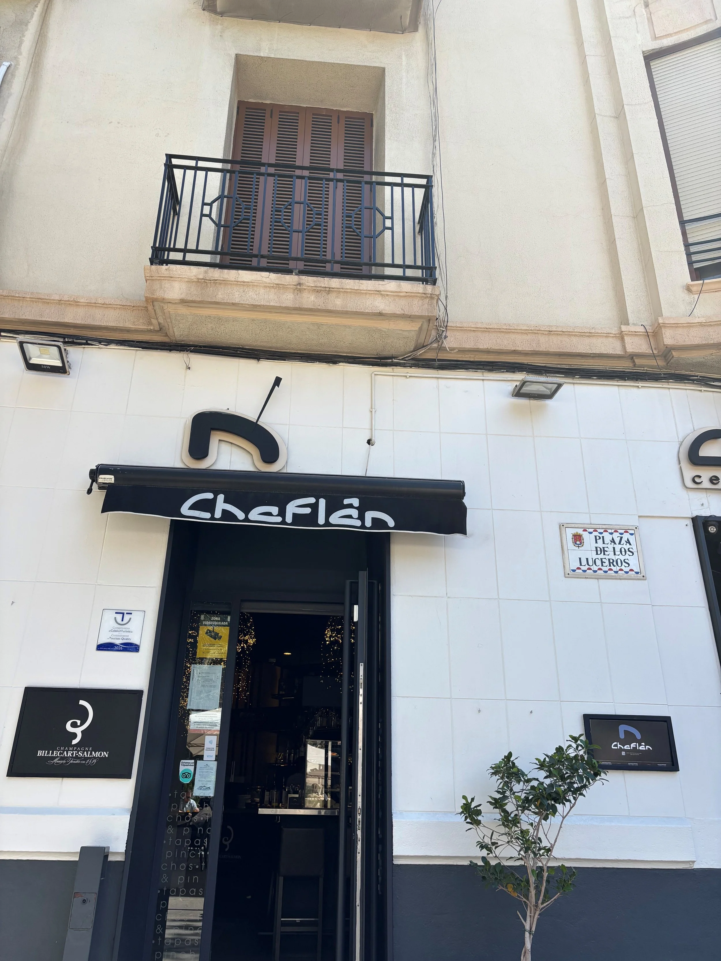 The image shows the entrance of a restaurant called Chefian, with a black door and signage displaying the name. There is a small tree in front of the building, two signs on the wall to the right of the door indicating awards or recognition, and a balcony above with closed shutters.