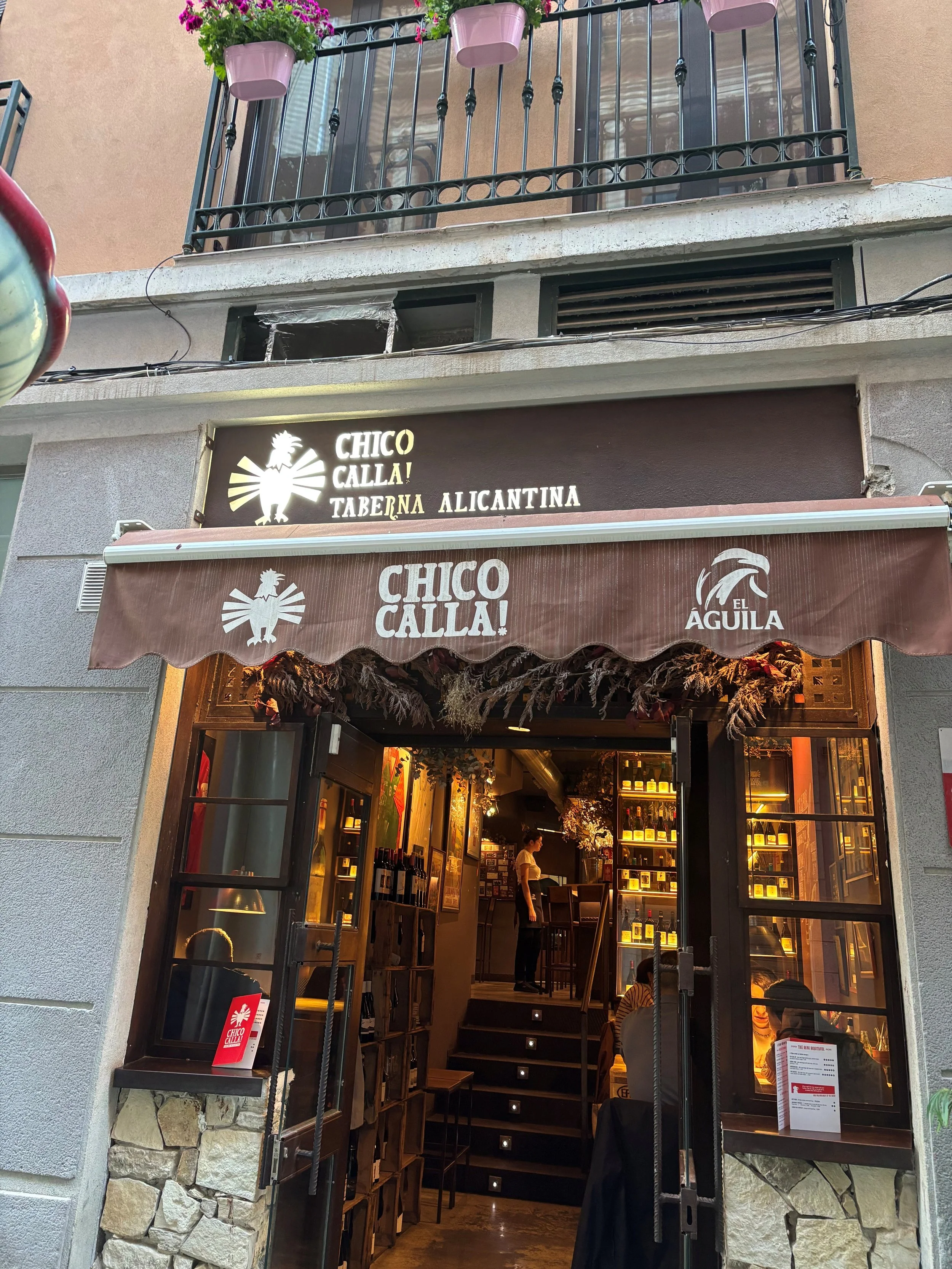 The exterior of a restaurant named Chico Calla with a sign that says 'Taberna Alicantina'. The entrance has open black-framed glass doors and a brown fabric awning with the restaurant's name and logo. Inside, warm lighting and a person can be seen at the counter, with shelves of wine bottles and decorated with dried flowers hanging from the ceiling.