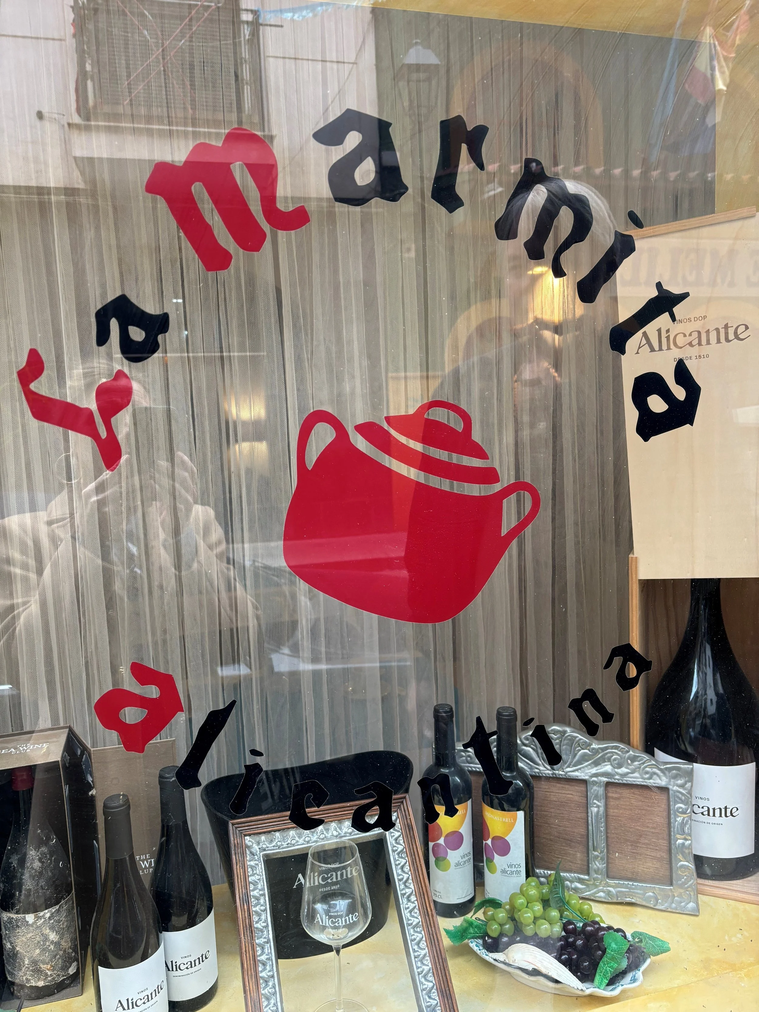 Window display with bottles of Alicante wine, a wine glass, grapes, a photo frame, and a sign with the text 'Alicante'.
