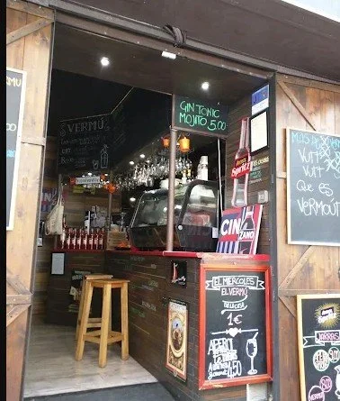 Small bar with a black chalkboard menu displaying gin tonic mojito for 5.00. Inside, a truck-shaped container holds bottles, with various signs and a wooden stool outside.