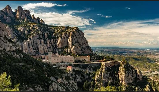 Scenic view of rocky mountains with a historic building at the base, overlooking a valley under a partly cloudy sky.