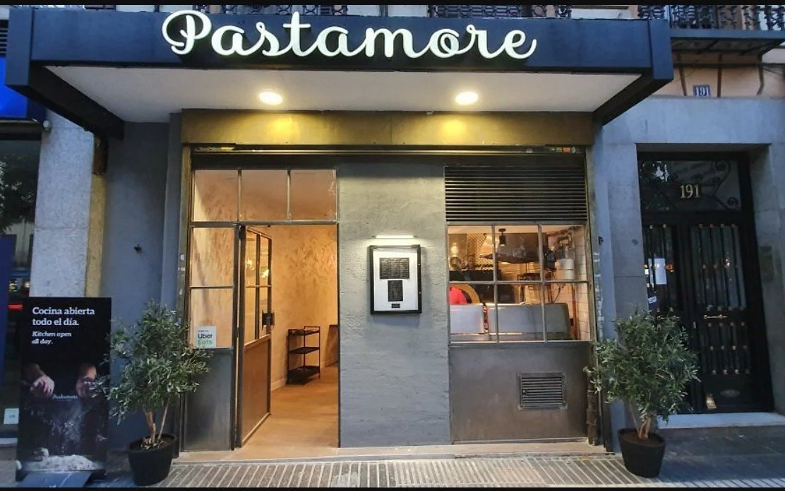 Front of a restaurant named Pastamore with a black and white sign, open entrance, and two potted plants outside.