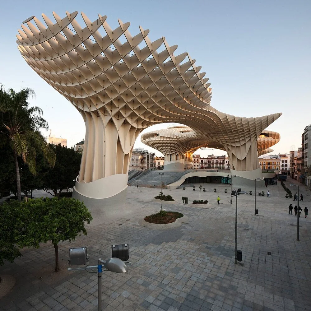 Unique modern architectural structure resembling large abstract trees with curving wooden lattice canopies, located in a spacious open plaza with some trees, benches, and a few people.
