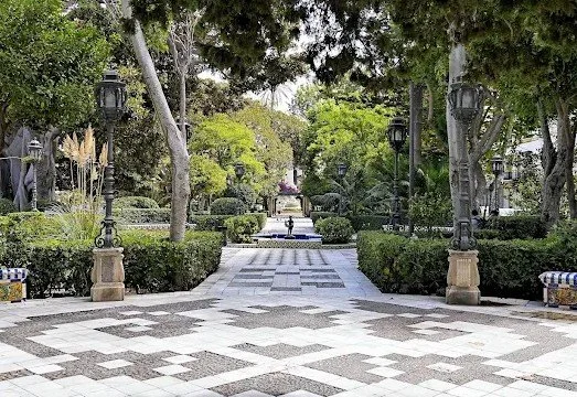 A paved walkway in a park with lampposts, trees, and greenery on both sides.