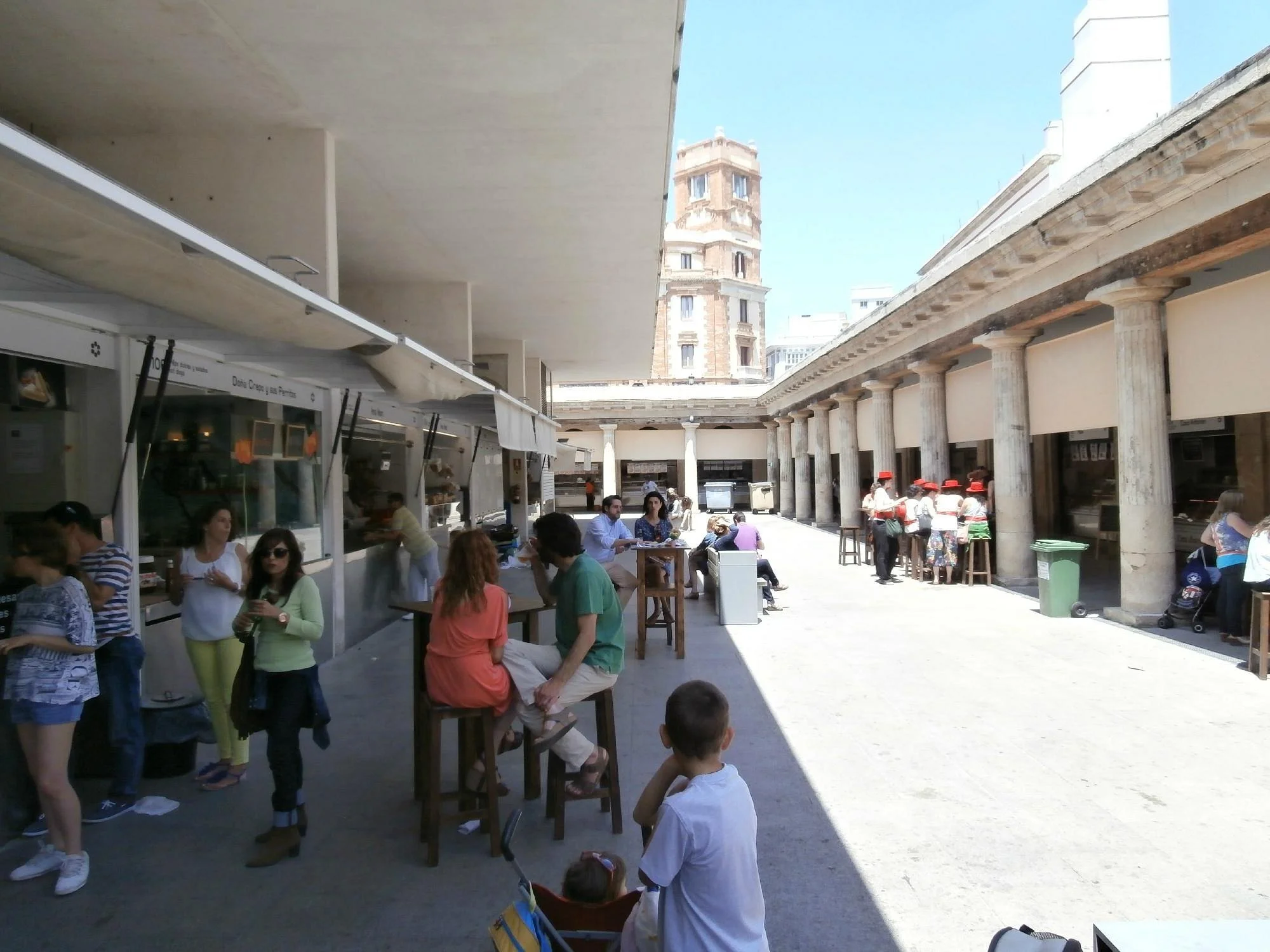 A busy outdoor marketplace with people sitting at wooden tables and standing around. The space is shaded by a large overhang, with a Roman-style columned building in the background. Some people are wearing hats and casual summer clothing, and there are food stalls along one side.