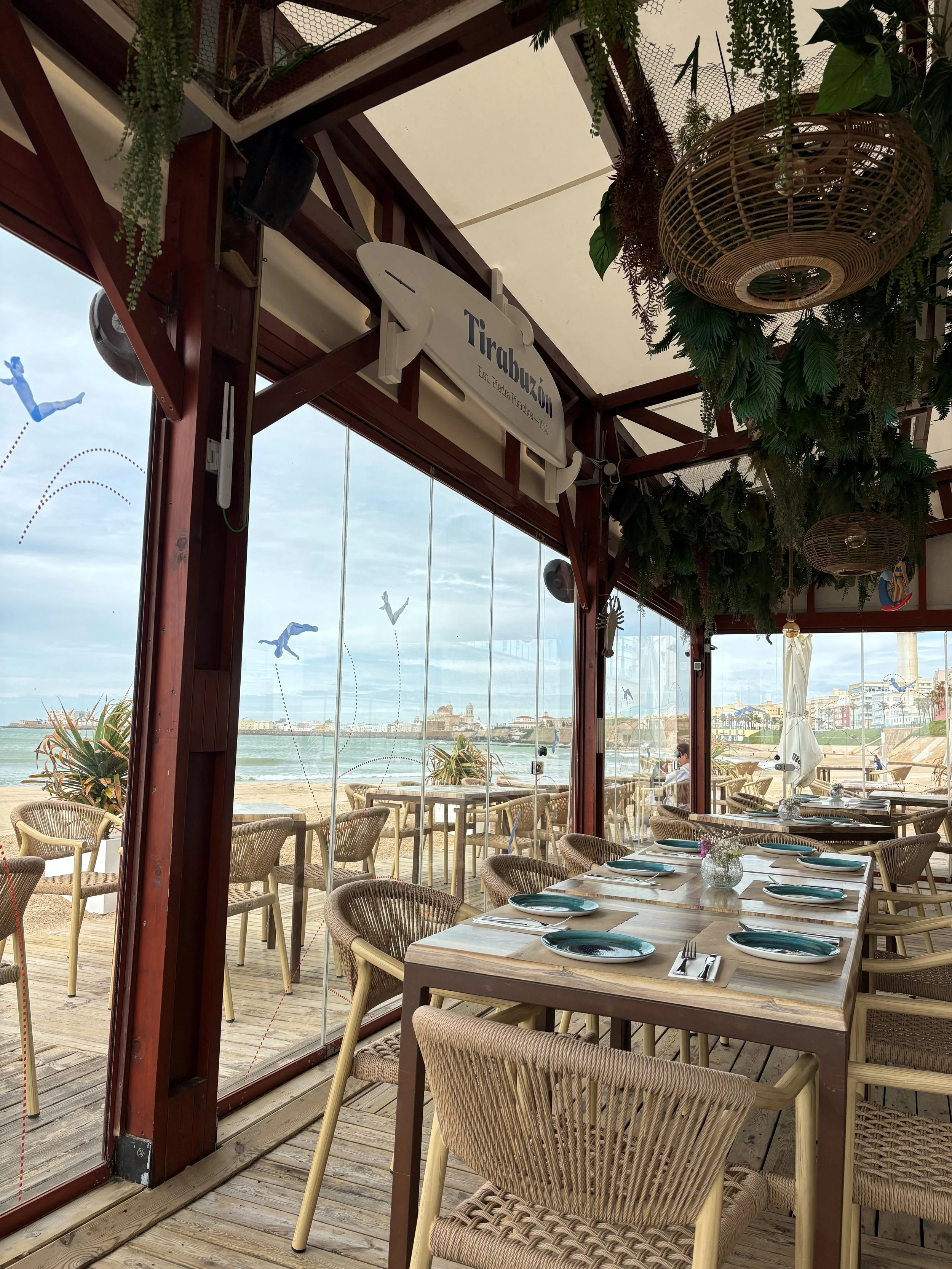 Indoor restaurant with wooden tables and wicker chairs, overlooking a beach with umbrellas, ocean, and buildings in the background. Decor includes hanging plants and wicker light fixtures.