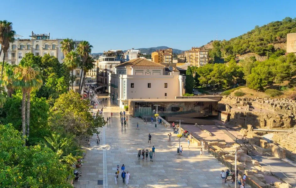 A busy plaza with people walking, surrounded by trees, buildings, and rocky ruins, under a clear blue sky.