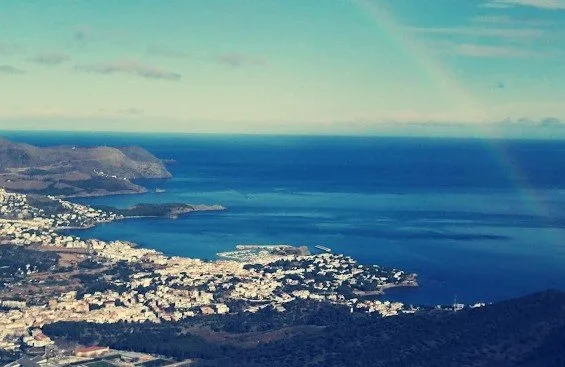 Aerial view of a coastal city with buildings, a bay, and blue ocean water, with mountains in the background and a partly cloudy sky.