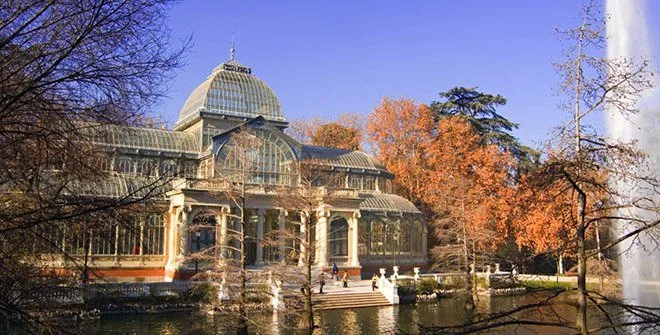 Glass greenhouse surrounded by trees with fall foliage, beside a pond with fountain in foreground.