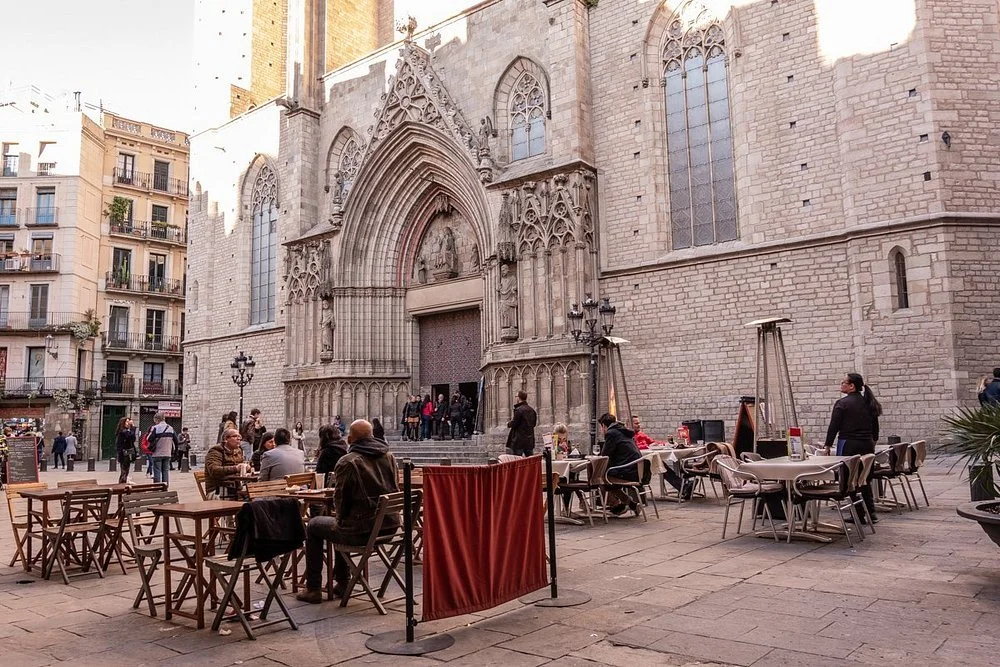 People sitting and standing outside a gothic-style cathedral, with outdoor tables and chairs, in an urban setting.
