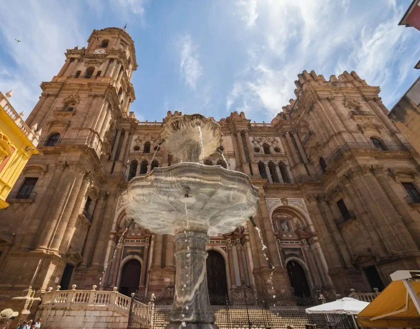 A historic cathedral with ornate stone architecture and multiple towers, viewed from below, with a fountain in the foreground and a partly cloudy sky overhead.
