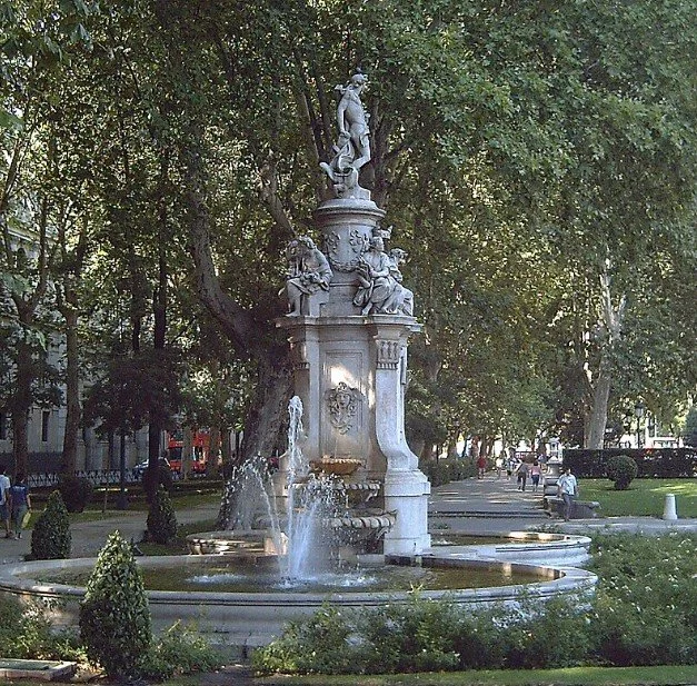 A historic ornate fountain with sculptures, water jets, and greenery in a park.