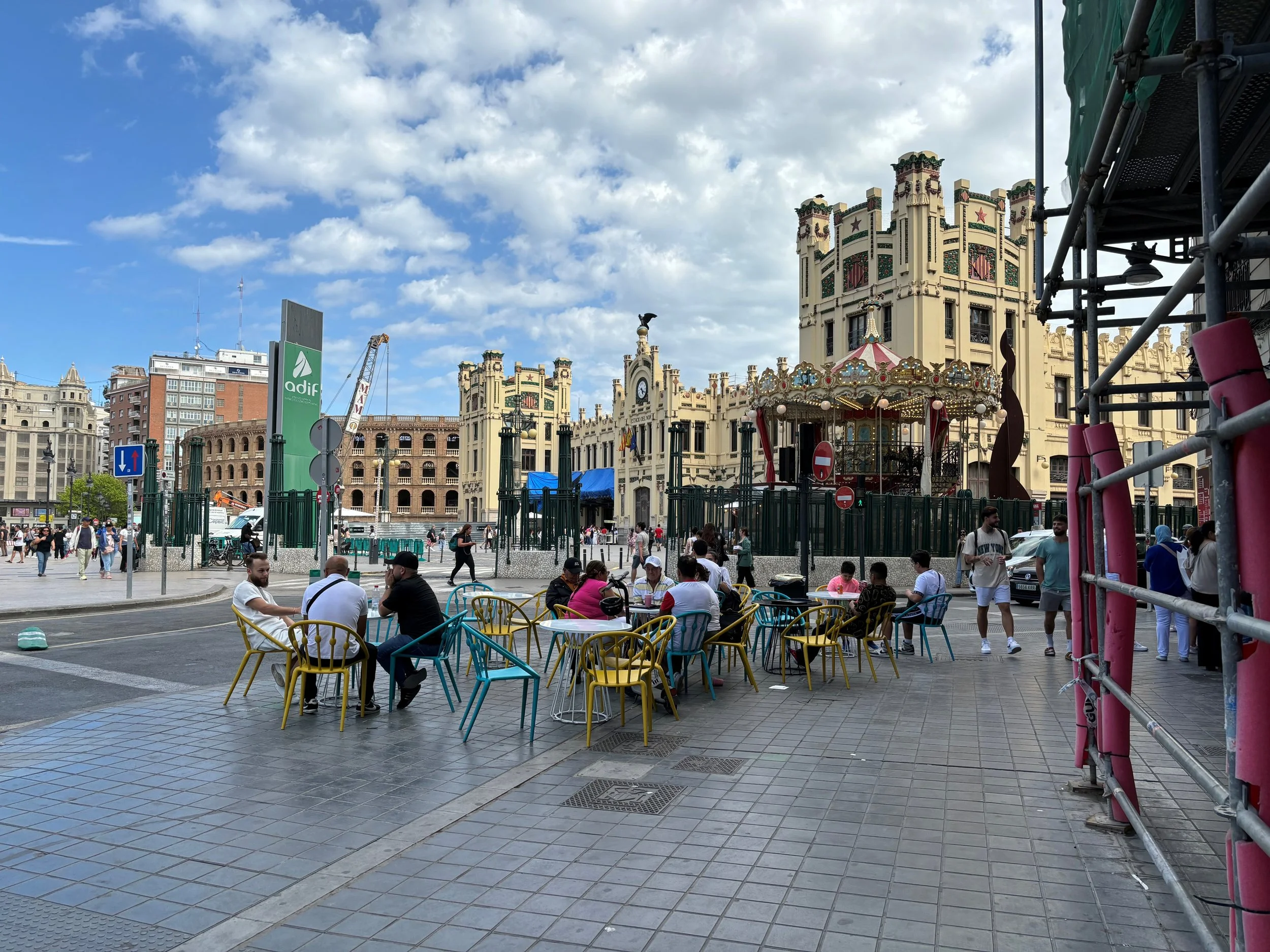 People sitting on colorful chairs and walking near a historic building with a carousel in front, under a partly cloudy sky.
