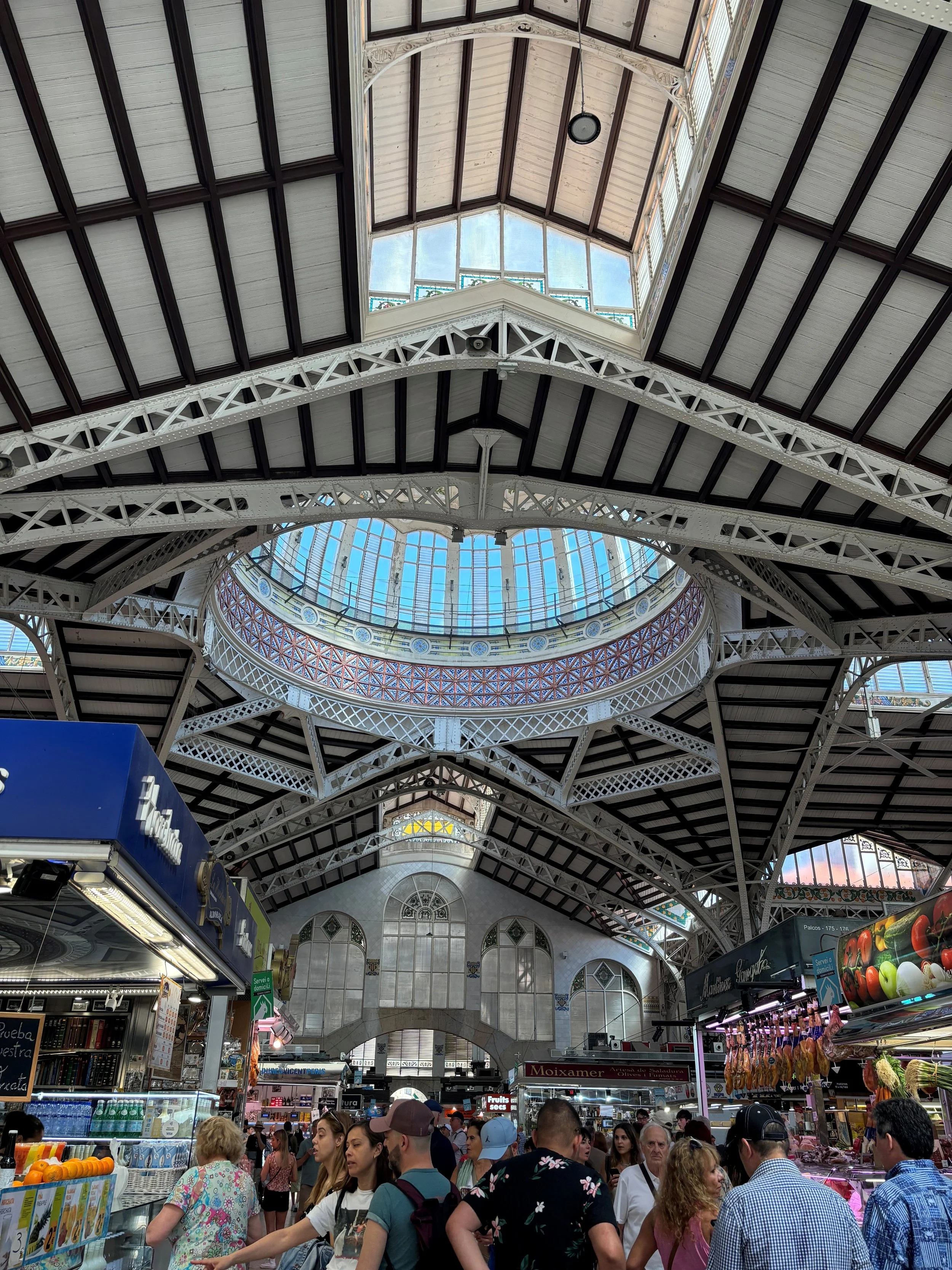 Interior view of a bustling marketplace with a high, glass-domed ceiling supported by white metal arches, filled with shoppers and vendors.