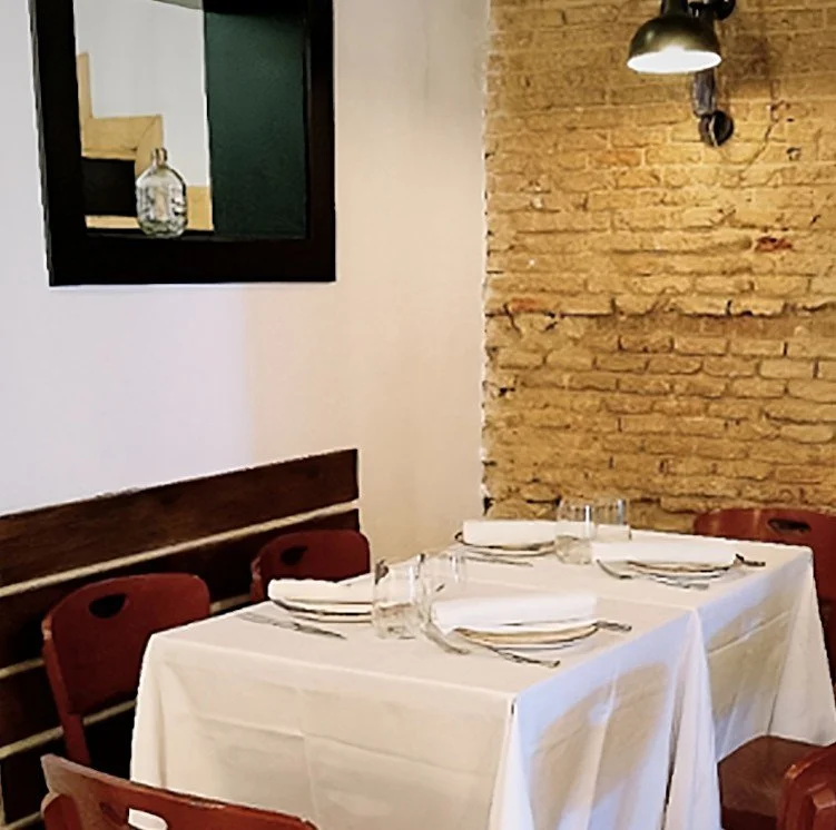 A dining table set with a white tablecloth, plates, napkins, and glasses in a restaurant with exposed brick wall and modern lighting.