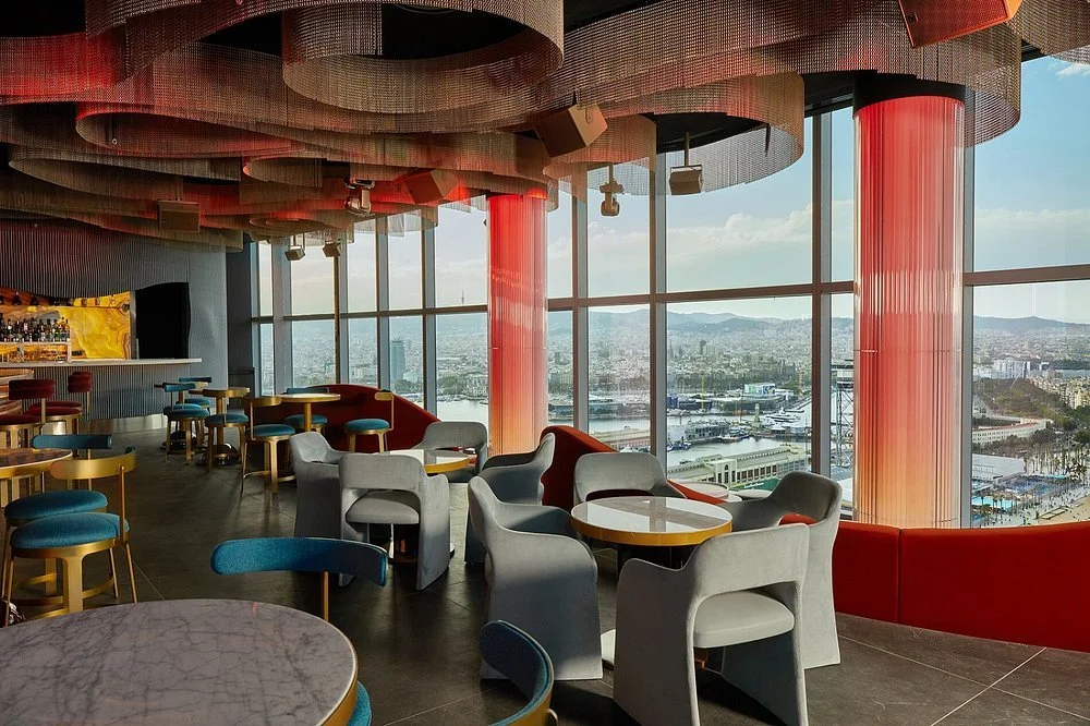 Empty modern lounge area with colorful chairs, round tables, large floor-to-ceiling windows with a cityscape view, and decorative ceiling panels.