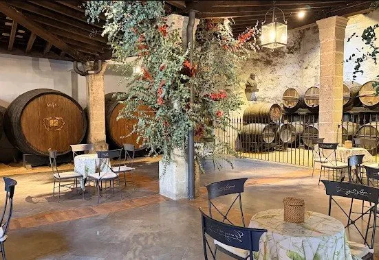 Wine barrels stored in an indoor wine cellar with rustic decor, surrounded by tables and chairs for tasting or dining.