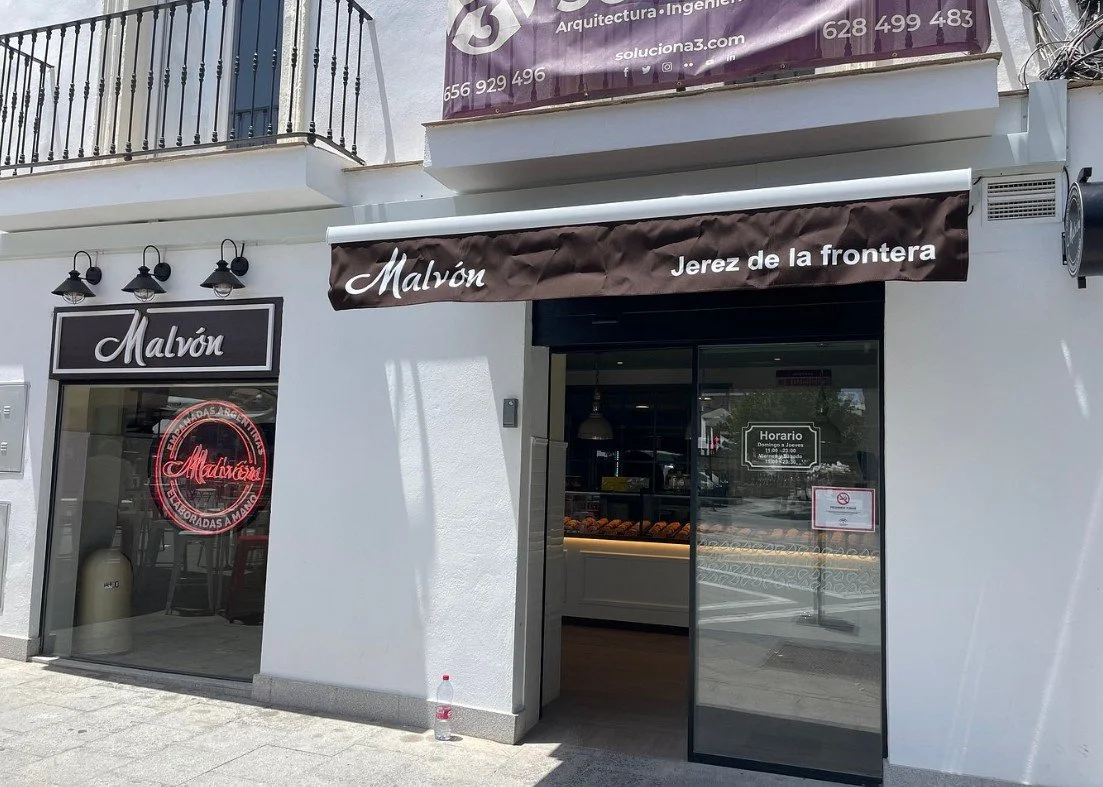 Front of a bakery shop named 'Malvón' with a sign indicating it is in Jerez de la Frontera. The shop has a glass door and window displaying baked goods inside. There is a brown awning above the entrance with the text 'Malvón Jerez de la frontera'. A neon sign inside the window reads 'Malvón' and 'Repostadas a mano'. Outside, a plastic water bottle is on the ground.