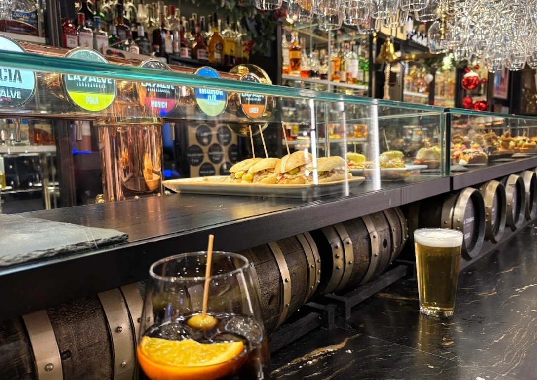 Bar with glass display case containing sandwiches, bottles of alcohol, and hanging wine glasses, with two drinks on the counter: a cocktail with orange slice and cherry, and a glass of beer.