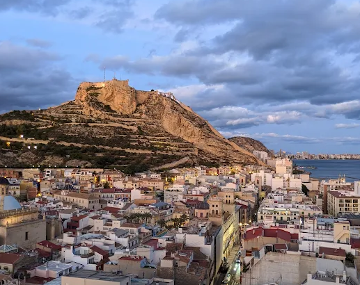 Cityscape of a coastal city with a large hill or small mountain in the background, under a partly cloudy sky.