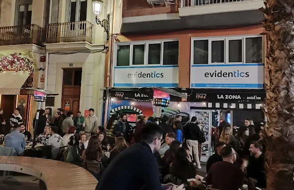 Nighttime outdoor scene with many people sitting at tables outside a bar or restaurant on a city street. Buildings with shops and signs, including 'Evidentics,' are visible in the background, along with a palm tree.