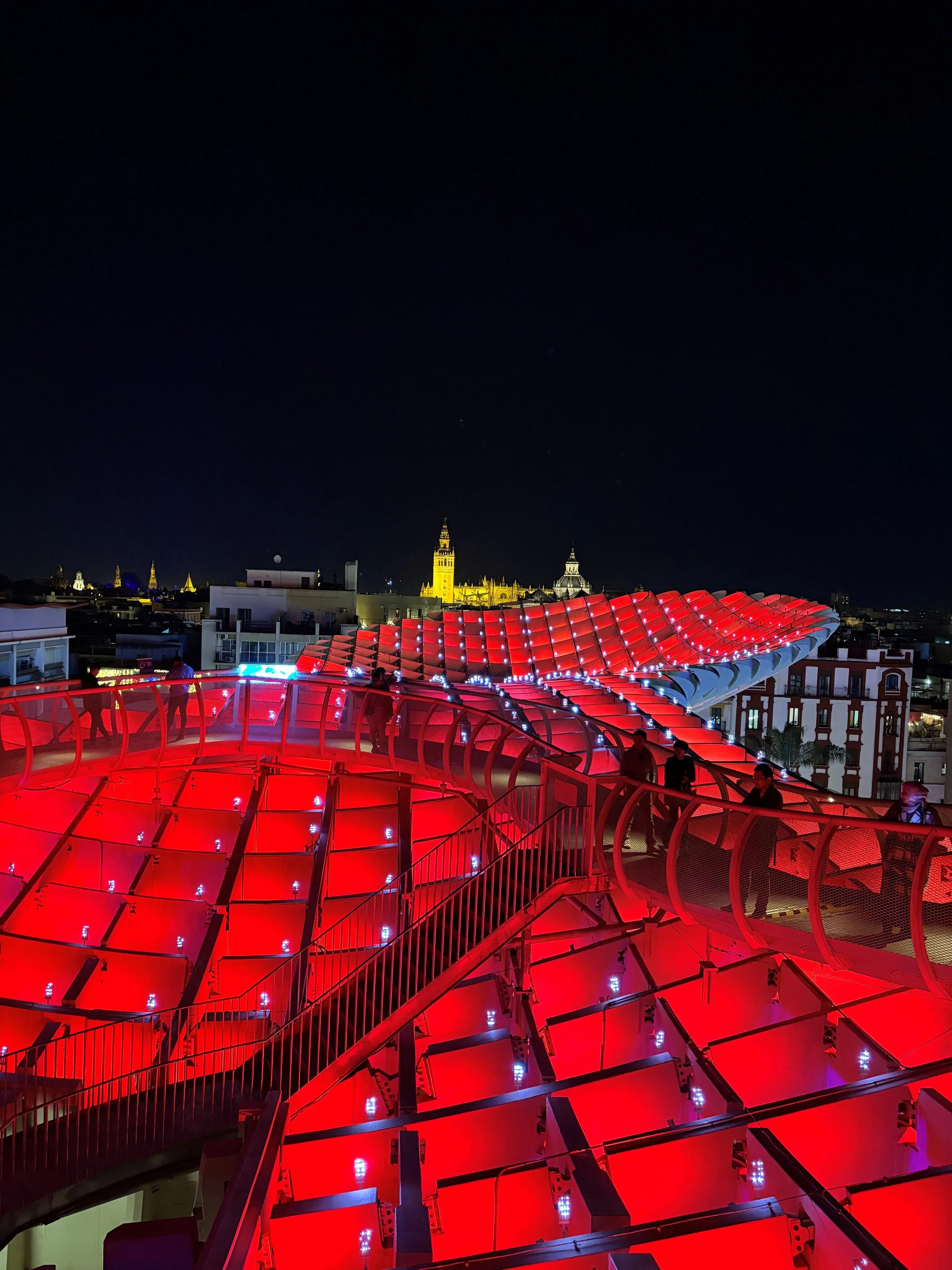 Night view of a city skyline with illuminated historic buildings and modern red-lit rooftop structure.