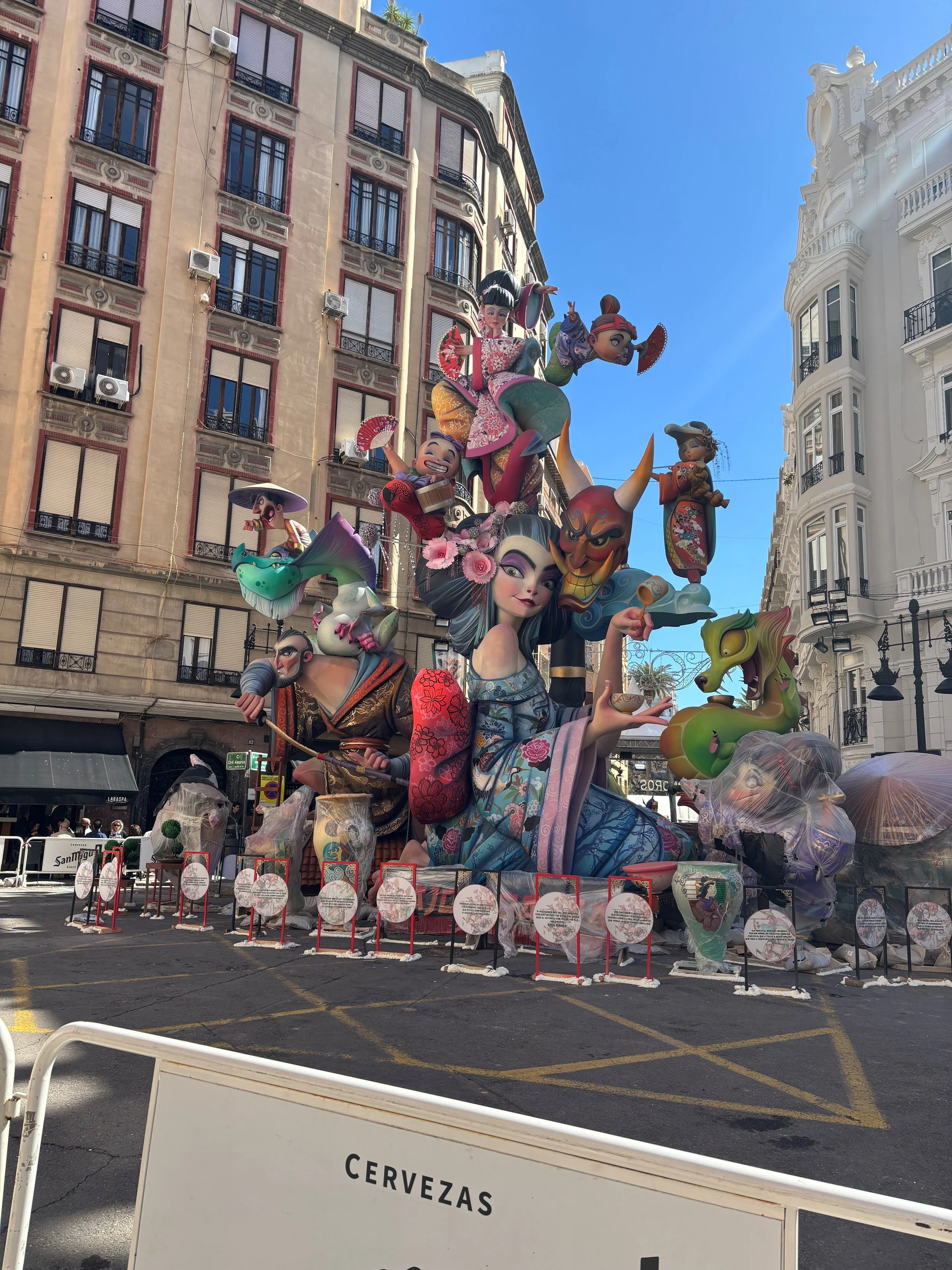 Colorful parade float with large, animated Asian-inspired characters and figures in traditional clothing, set against urban buildings under a clear blue sky.