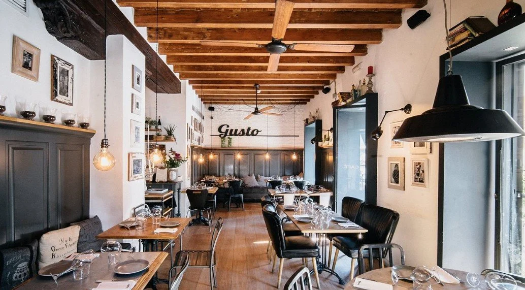 Interior of a restaurant with wooden ceiling beams, dark gray wainscoting, modern hanging pendant lights, framed photos on the white walls, and tables set with glassware and napkins. There are black and metal chairs, and a sofa with cushions at the back, with a sign reading "Gusto" on the wall.