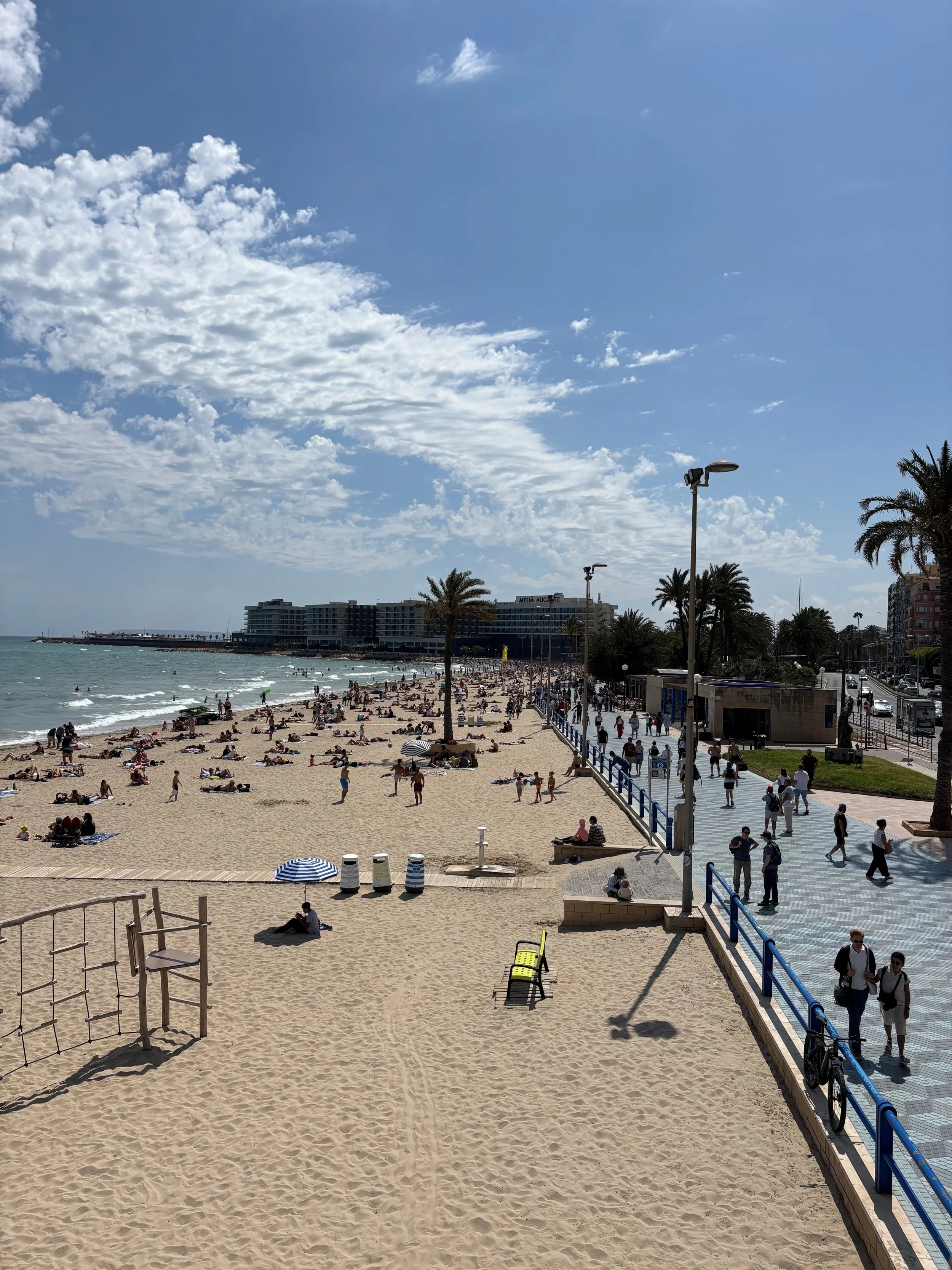 A busy beach scene with many people relaxing on the sandy beach, some under umbrellas, and others walking along the shoreline. The ocean is visible with gentle waves, and there are buildings, palm trees, and a boardwalk with pedestrians and cyclists in the background. The sky is partly cloudy with some scattered clouds.