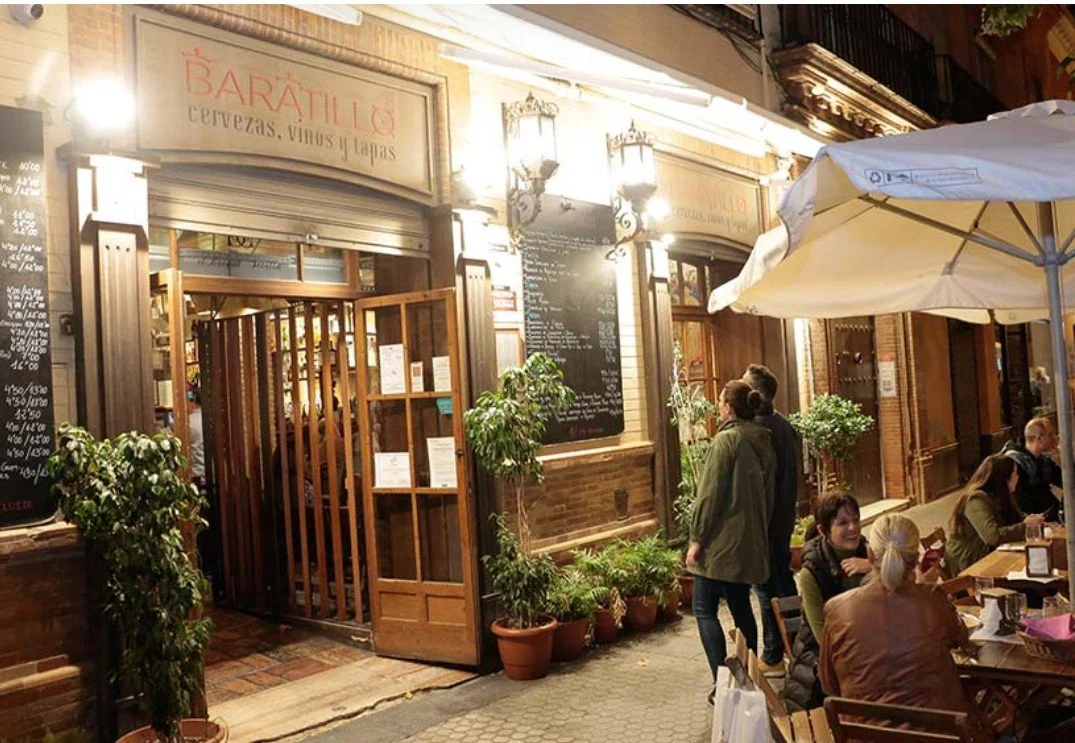 Exterior view of a restaurant with outdoor seating, potted plants, and people dining under an umbrella at night.