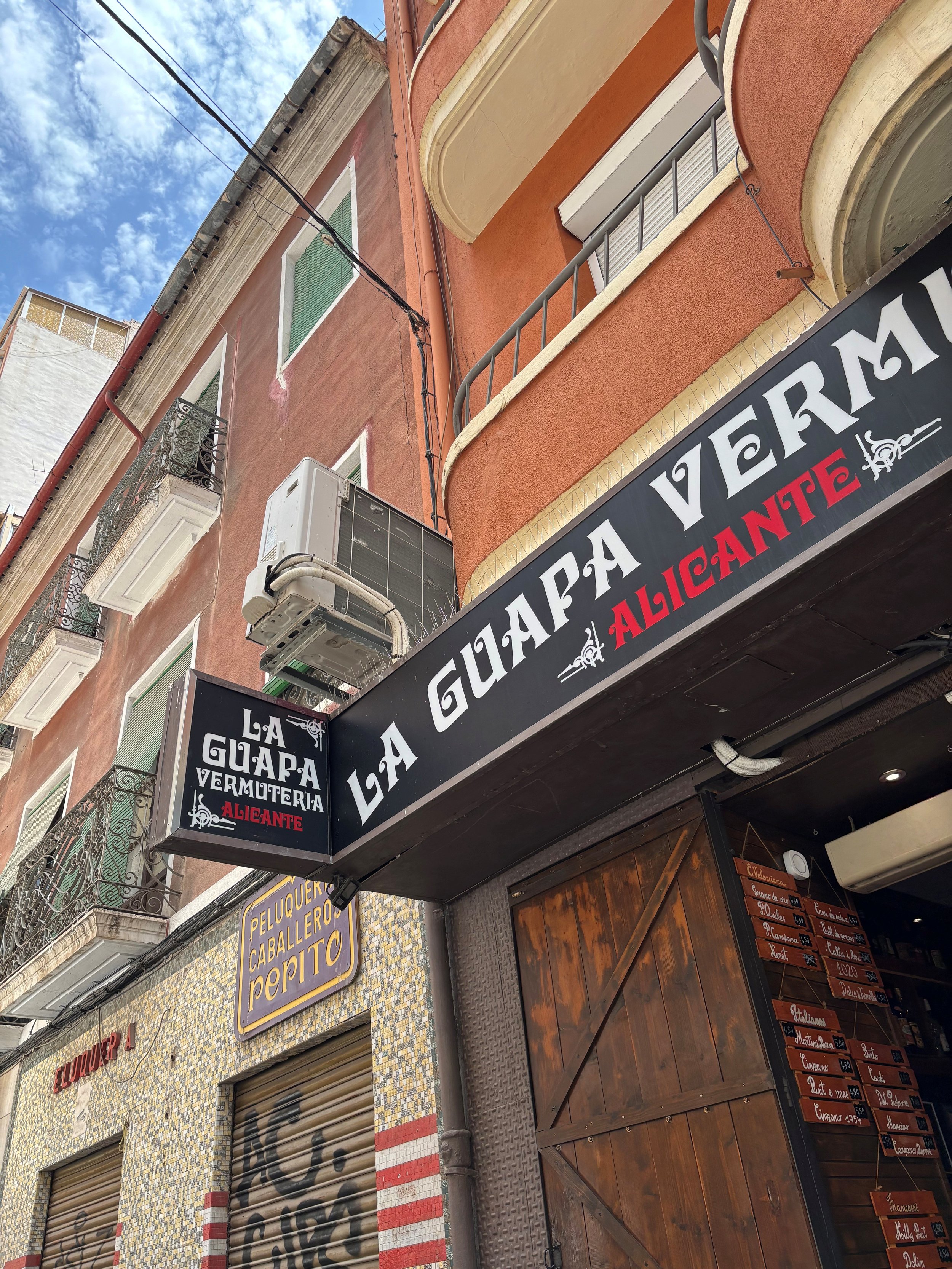 Street view of a building with a sign for La Guapa Vermuteria Alicante. Metal shutters on some storefronts, upper windows with small balconies, an air conditioning unit, and wiring along the wall. The sky is partly cloudy.