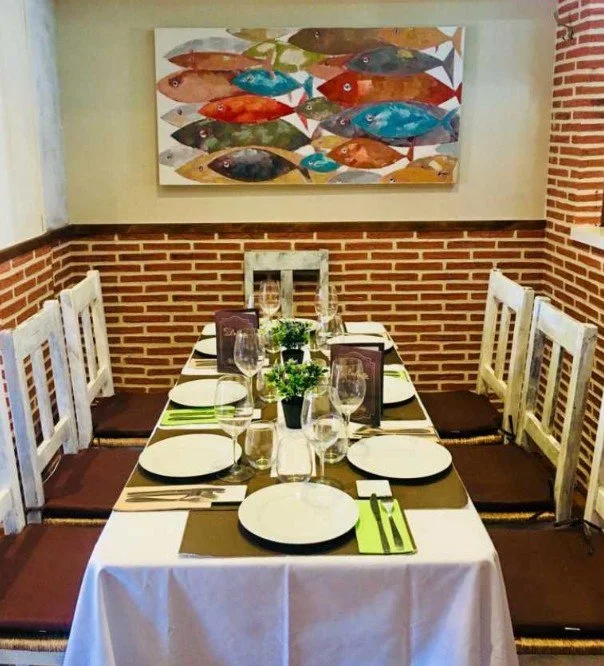 Table set for six with white tablecloth, plates, green utensils, glasses, and small plants, in a room with brick walls and a colorful fish painting.