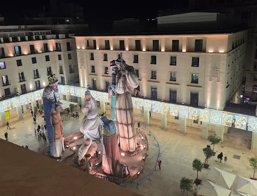 Night view of a city square with large illuminated statues depicting two kings or wise men, decorated with crowns and royal clothing, with a large building in the background.